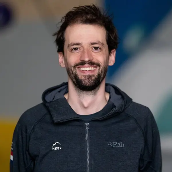 Een man met kort bruin haar en een baard staat binnen, gekleed in een zwart Rab-T-shirt en met een lanyard om zijn nek. Achter hem staan mensen in identieke shirts met het opschrift NED, en boven hun